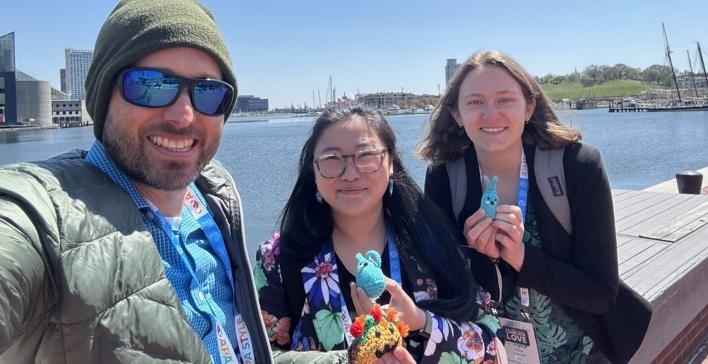 Todd Ruecker, Hannah, and Olivia smile and hold up their creatures. Baltimore's Inner Harbor is in the background.