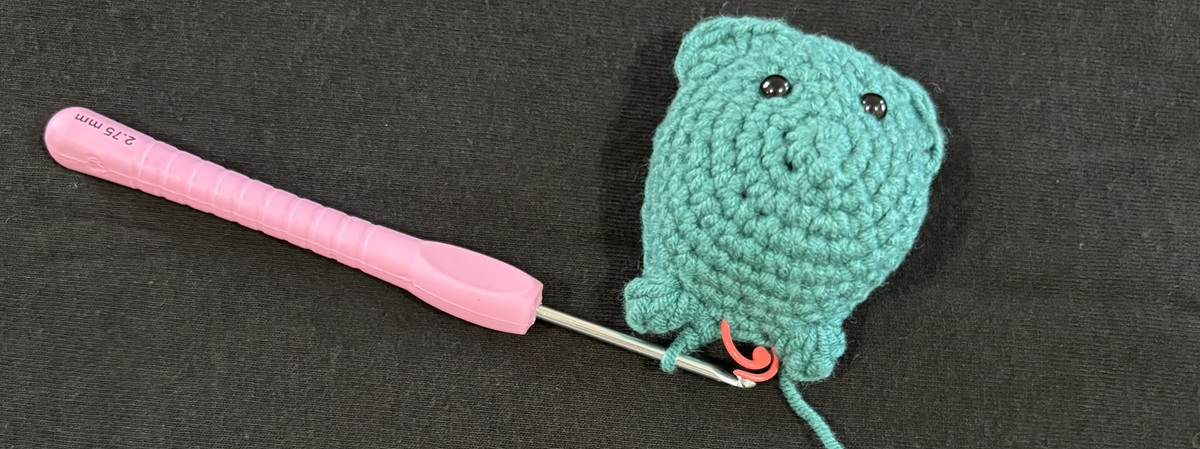 Banner The front half of a blue crochet cat attached to a purple crochet hook on a black background.