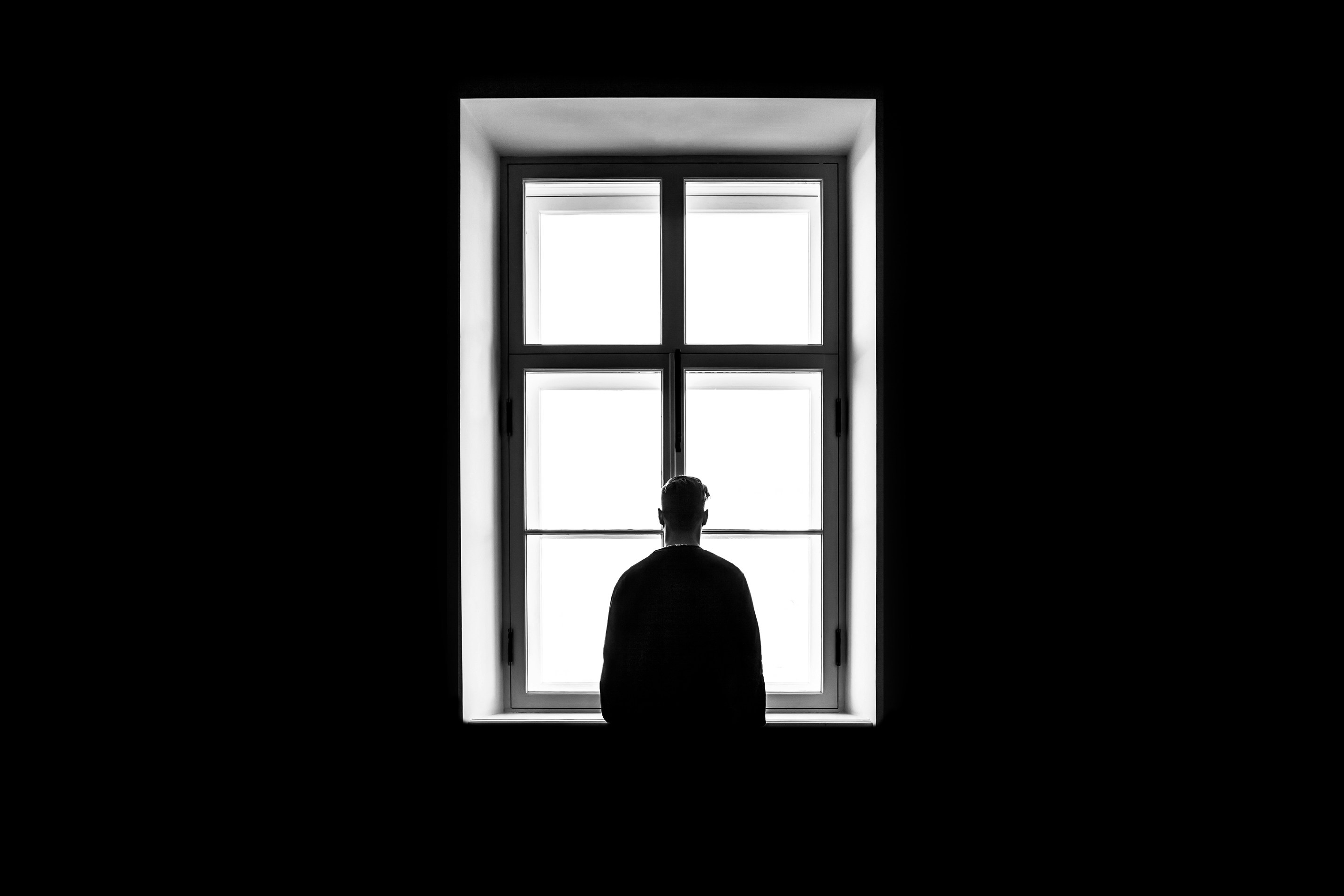The silhouette of a man standing solitary in front of a large window.