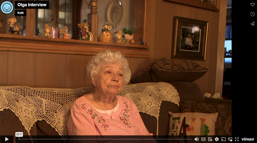 Olga, an elderly lady with white hair and a pink sweater, sits on a couch looking up into the distance