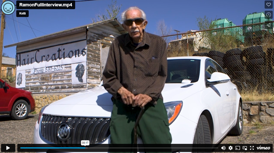 Ramon, an elderly man in sunglasses, leans against a white Buick sedan. He's wearing a brown button-up shirt and green slacks. He holds a cane in his hands. Behind him is a shed with an advertisement painted on the side of it that says Hair Creations. The sun casts hard shadows on his face and the sign