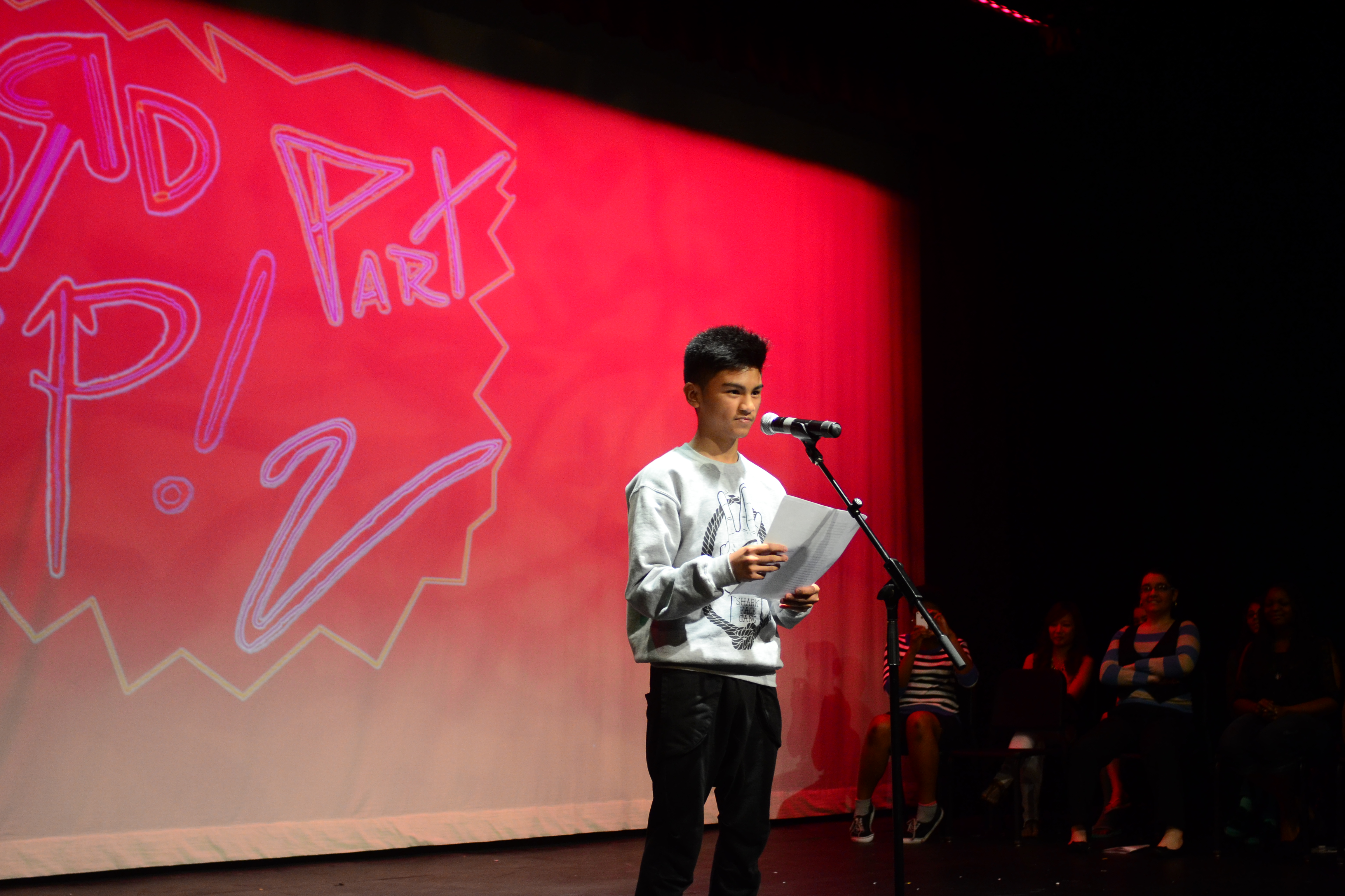 A person with short dark hair wearing a sweatshirt and jeans stands on stage in front of a bright red screen reading at a microphone