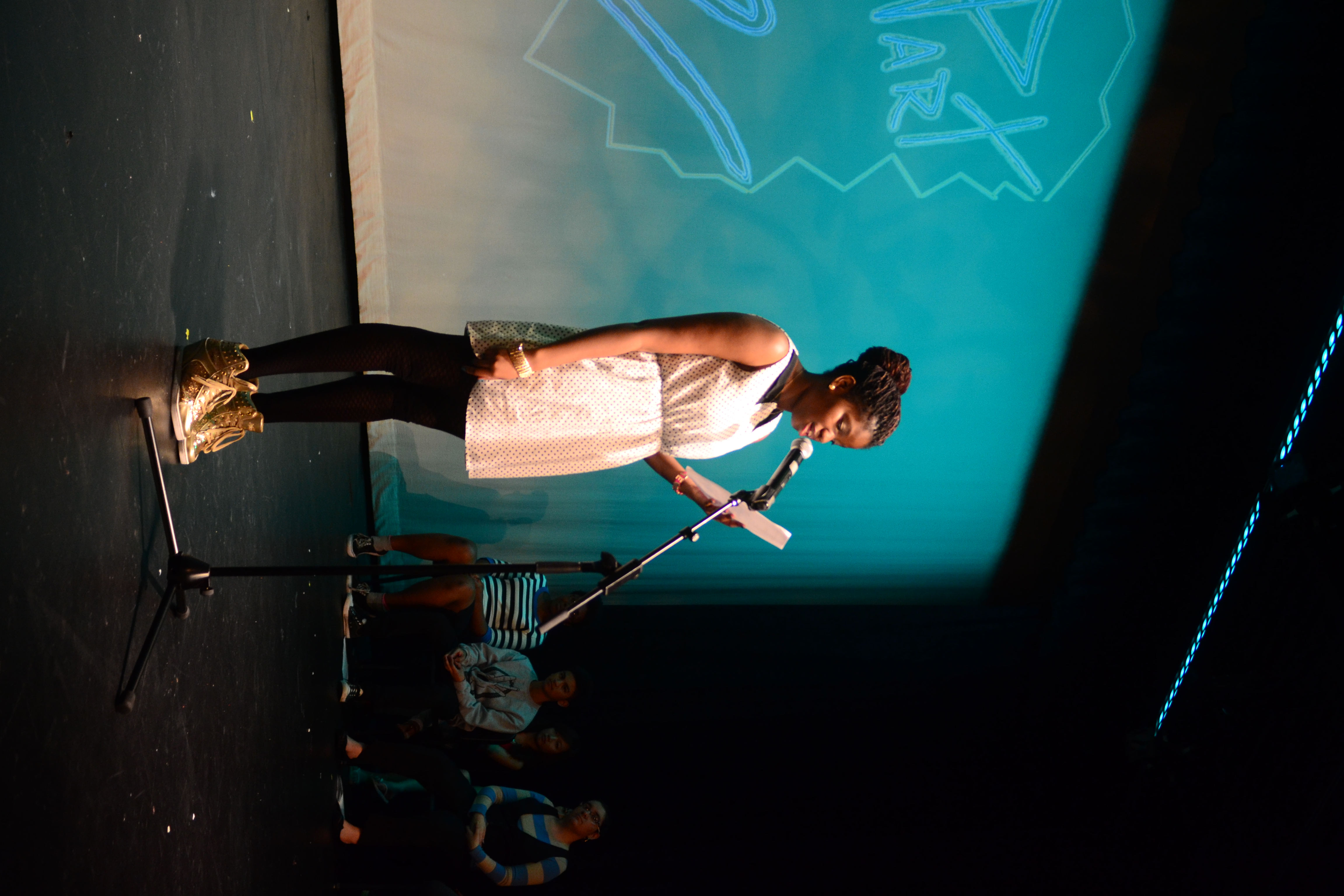 A person with dark hair in a bun and wearing a white polkadot dress, black leggings, and gold hightops performs on stage at a microphone.